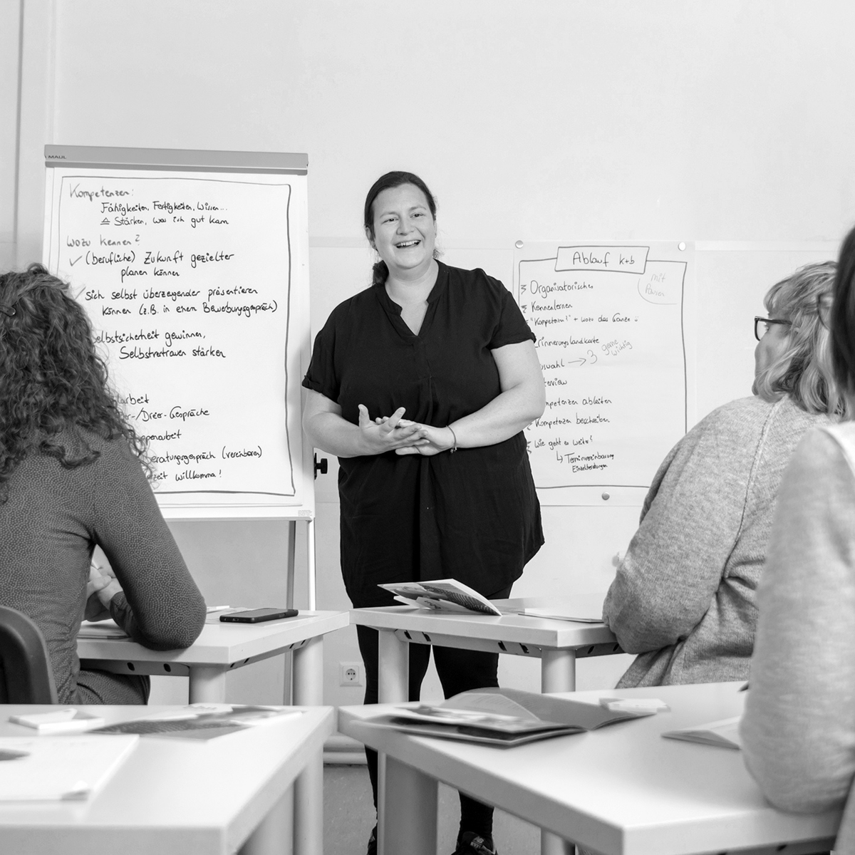 Eine Frau steht vor einem Flipchart und spricht zu einer Gruppe von Personen, die an Tischen sitzen und Unterlagen vor sich haben.