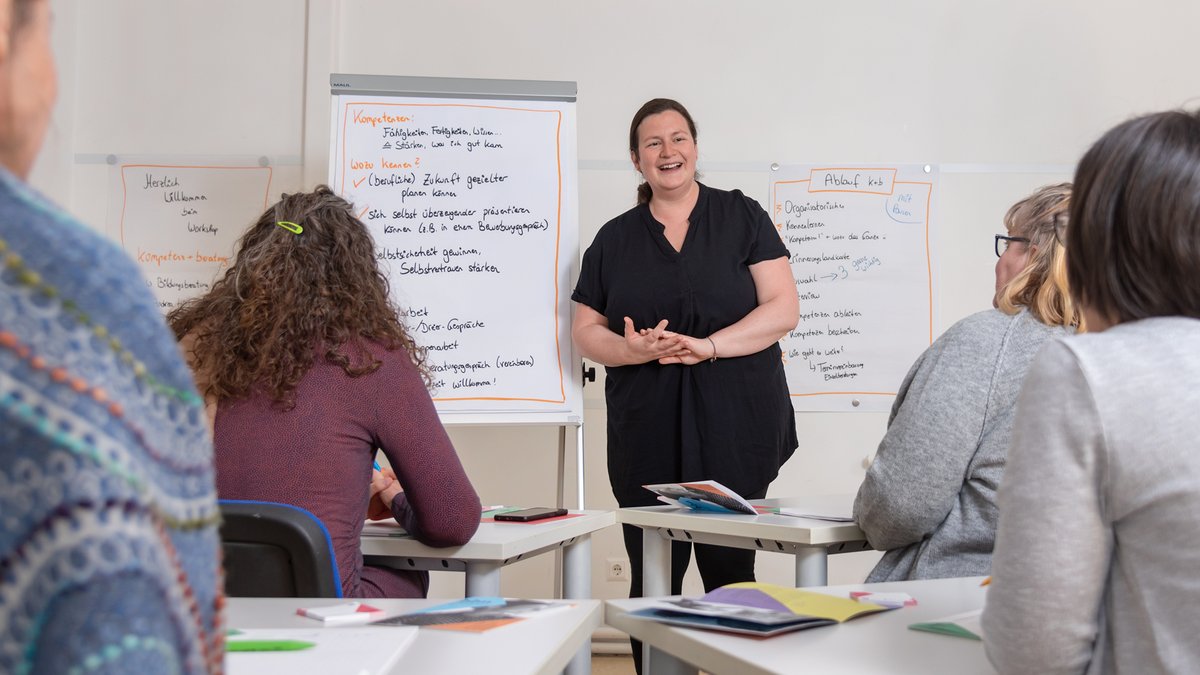 Eine Frau steht vor einem Flipchart und spricht zu einer Gruppe von Personen, die an Tischen sitzen und Unterlagen vor sich haben.